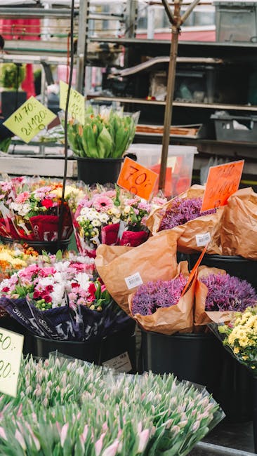 A vibrant display of fresh flowers arranged in black plastic buckets at an outdoor market stall, featuring a variety of floral arrangements including pink and white chrysanthemums, purple lavender, pi