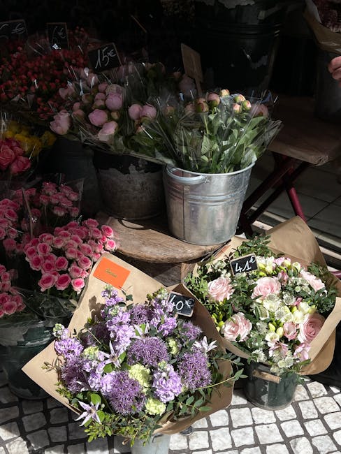 A display of fresh flower bouquets at a flower shop, arranged on a rustic wooden table. The arrangements include a bouquet of light pink roses with green foliage wrapped in brown craft paper, a purple
