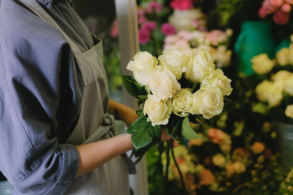A person dressed in a grey apron is holding a bouquet of fresh, creamy white roses with lush green leaves. The roses are arranged tightly together, displaying full, layered petals, and are wrapped in 