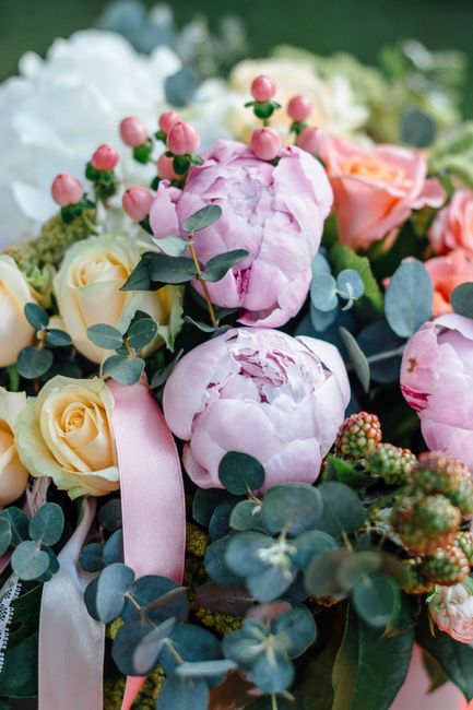 A close-up view of a floral arrangement featuring soft pink peonies with lush, ruffled petals, complemented by pale yellow roses exhibiting smooth, spiraled blooms. The bouquet is accented with small 