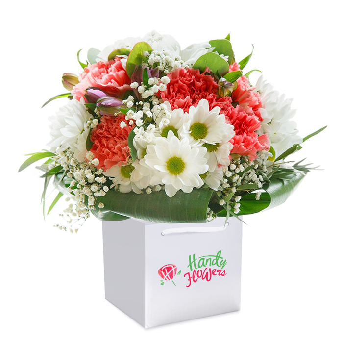 A sophisticated and vibrant floral display is arranged upright within a crisp white gift bag, its contemporary silhouette highlighted by a seamless white backdrop-a scene that captures both modern style and the timeless charm of Kingsbury. The bouquet features an artful blend of coral-pink, peach, and radiant red carnations, their frilled petals creating a multidimensional texture that feels as luxurious as it looks. Prominently nestled between these lush blooms are pure white, daisy-like flowers with cheerful lime-green centers, adding a note of brightness and joie de vivre reminiscent of playgrounds in Roe Green or the gardens along Church Lane. The arrangement is interwoven with delicate sprays of baby's breath, whose airy white clusters impart a feather-light softness throughout. Veiled within the bouquet, hints of purple blush from closed buds lend intrigue, signalling future colour and floral evolution. Encircling the base, broad and shiny deep green foliage frames the entire piece, creating a supportive structure and a vivid contrast. The square white gift bag boasts strong ribbon handles, ready for efficient delivery to homes and offices across the Kingsbury area. This thoughtfully crafted bouquet is an ideal symbol of celebration or gratitude, designed with local expertise by Florist Kingsbury-adds an uplifting touch to birthdays, anniversaries, or heartfelt everyday occasions within the friendly neighbourhood.